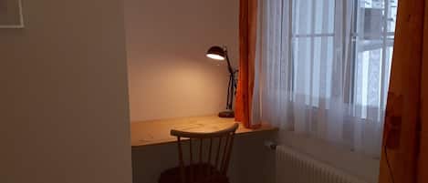Een kleine studeerkamer met een bureau, stoel, lamp en een raam met gordijnen.