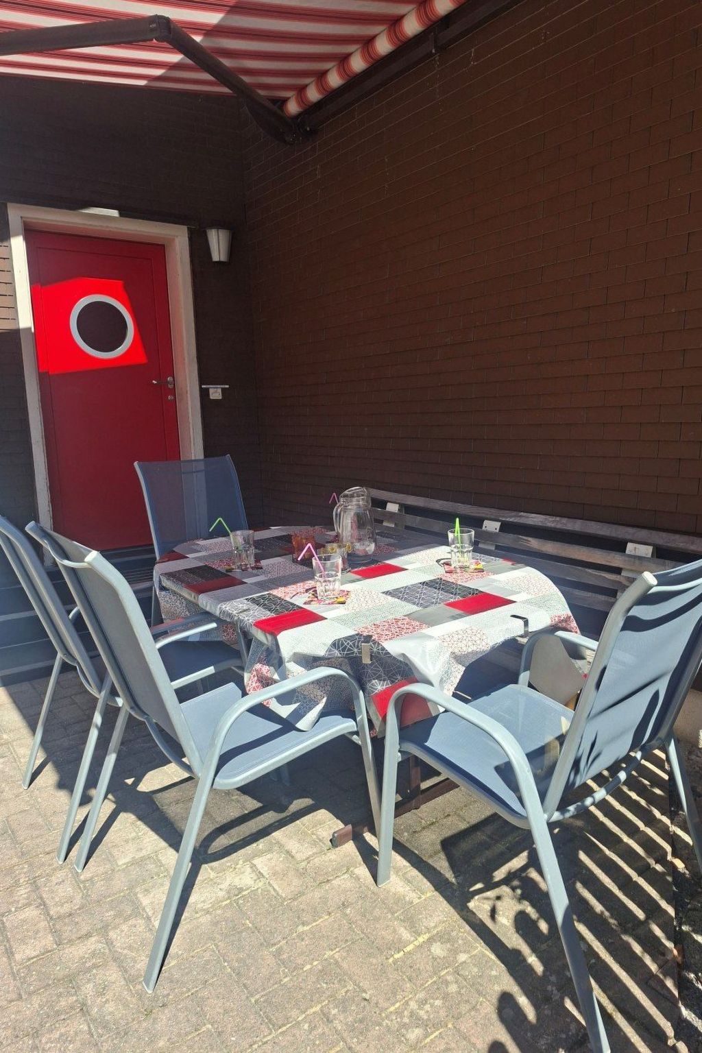 Un patio al aire libre con una mesa para cuatro personas, rodeado de sillas de metal, frente a un muro de ladrillo con una puerta roja.