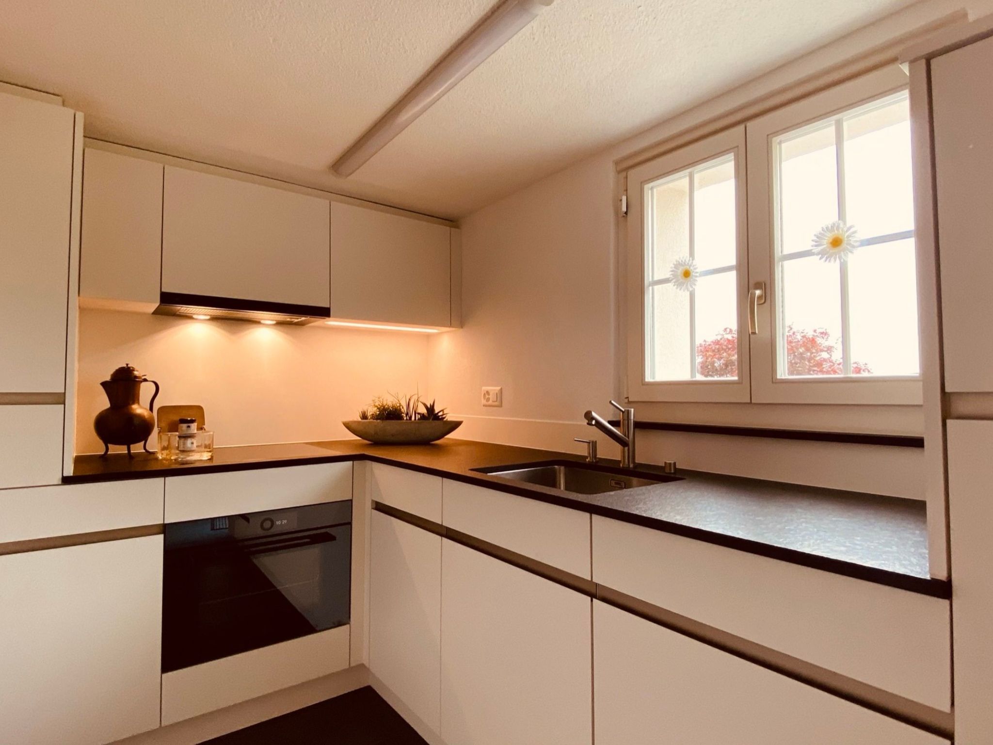 A modern kitchen with white cabinets, a black countertop, a built-in oven, and a window with flowers.