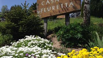 Ein hölzernes Schild mit der Aufschrift 'CASA FLURETTA', umgeben von einem Garten mit verschiedenen Blumen.