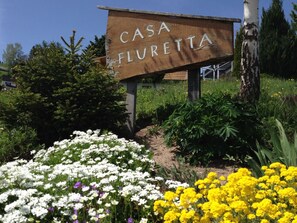 Ein hölzernes Schild mit der Aufschrift 'CASA FLURETTA', umgeben von einem Garten mit verschiedenen Blumen.