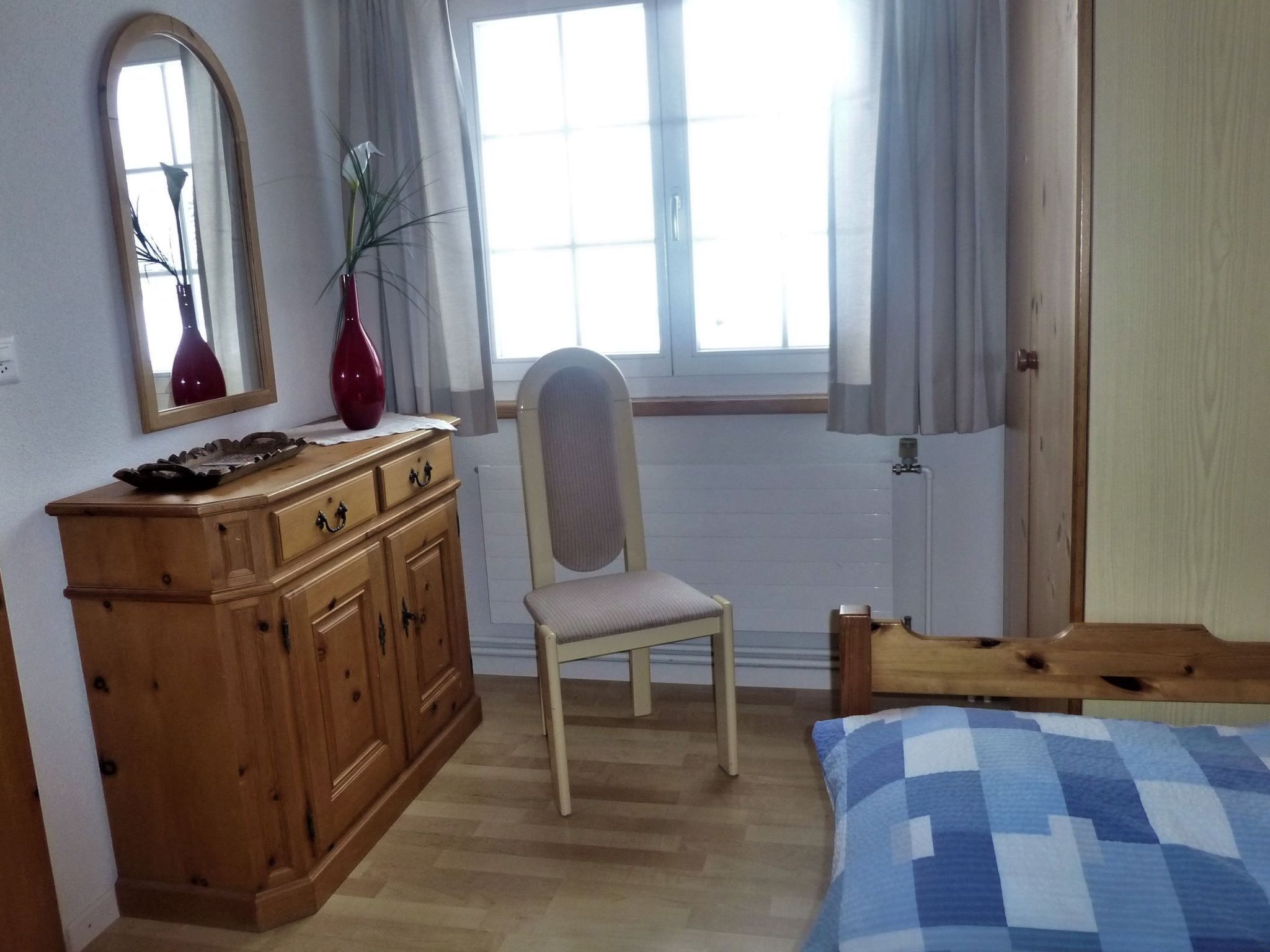 Ein Schlafzimmer mit einem Holzkapfen, einem Sessel, einem Bett mit blau-weiß kariertem Überwurf, einem Fenster mit Vorhängen und einem Spiegel an der Wand.