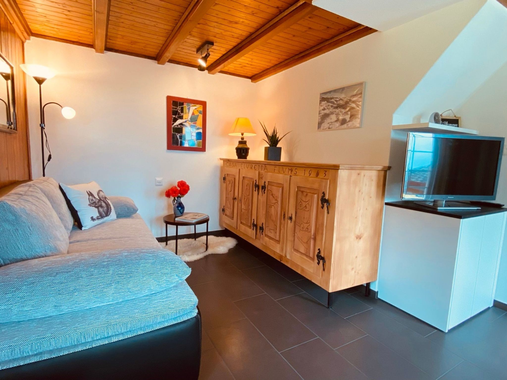 A cozy living room with a sofa, a small side table with a lamp, a cabinet with numbered doors, and a television on a stand.