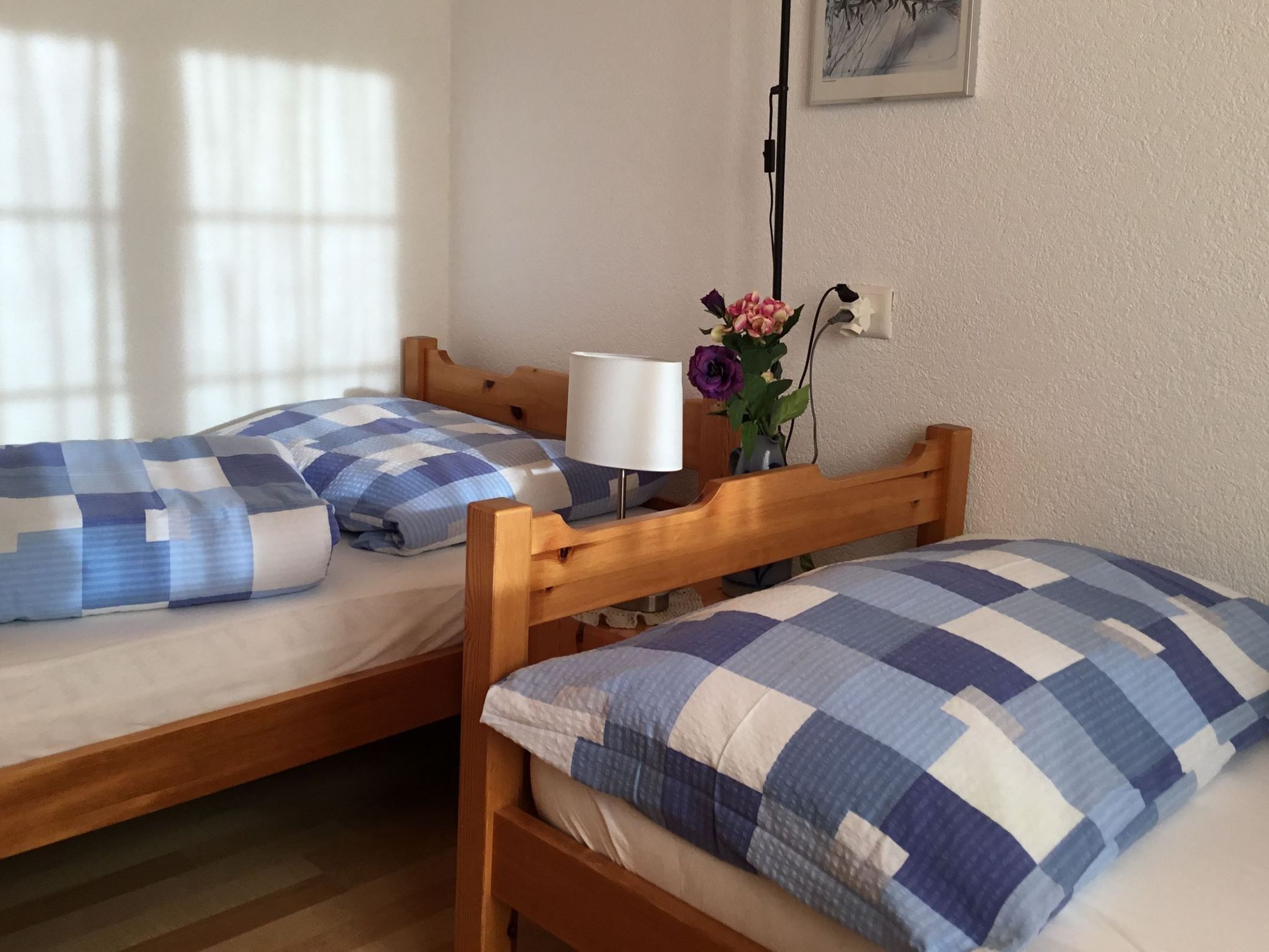Zwei Einzelbetten mit karierter Bettwäsche, eine Nachttischlampe, eine Vase mit Blumen und ein Bild an der Wand.