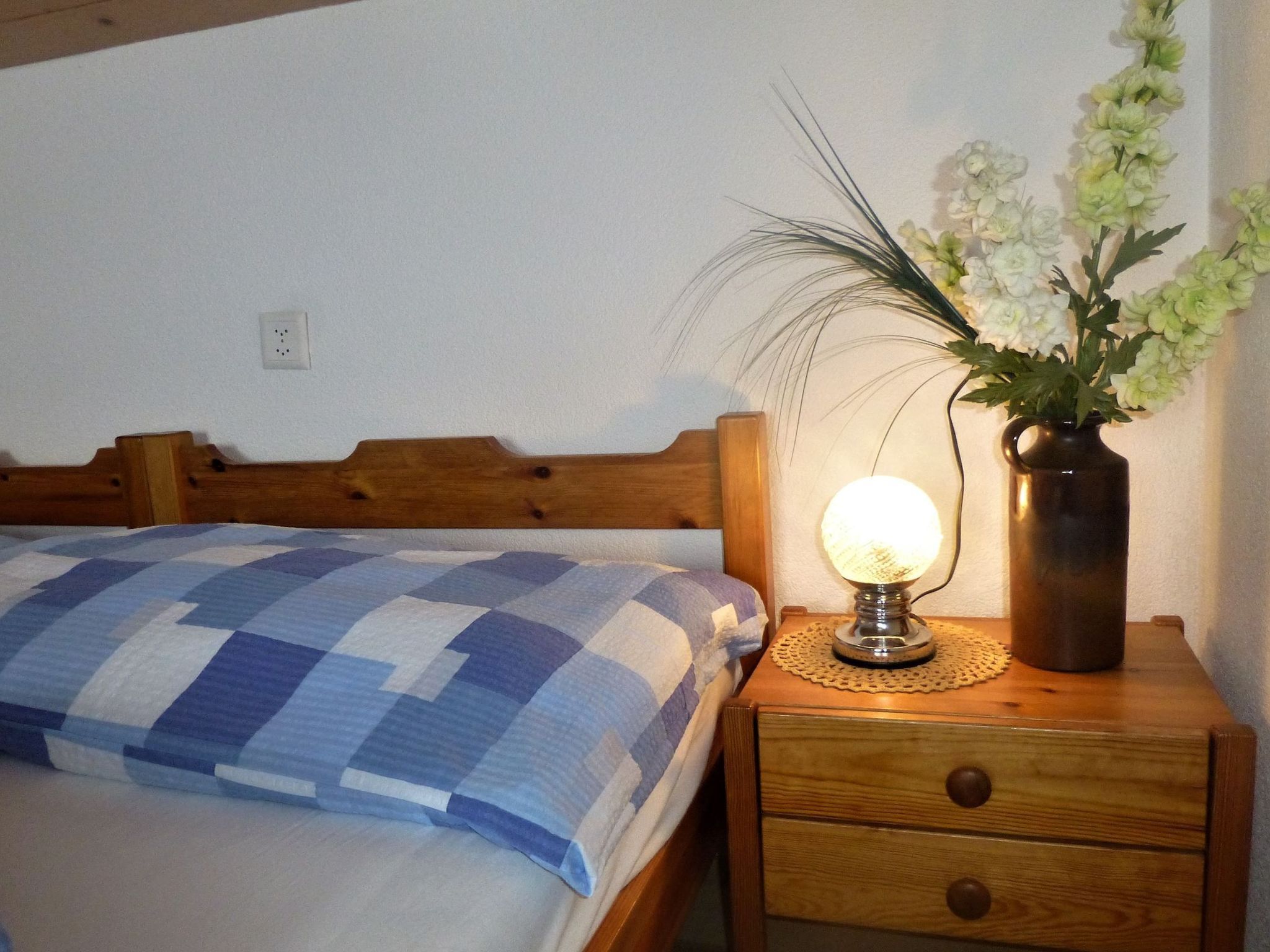 En seng med et ternet tæppe, en sengetavlelampe og en vase med blomster på et trænatbord.