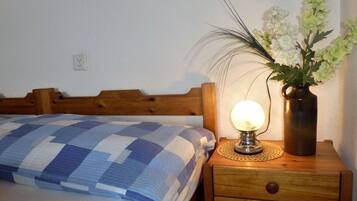 En seng med et ternet tæppe, en sengetavlelampe og en vase med blomster på et trænatbord.