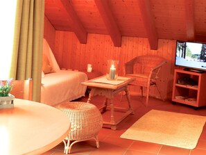 Une pièce confortable avec un lit, un fauteuil en osier, une petite table avec une bougie, un téléviseur et un pouf en osier.