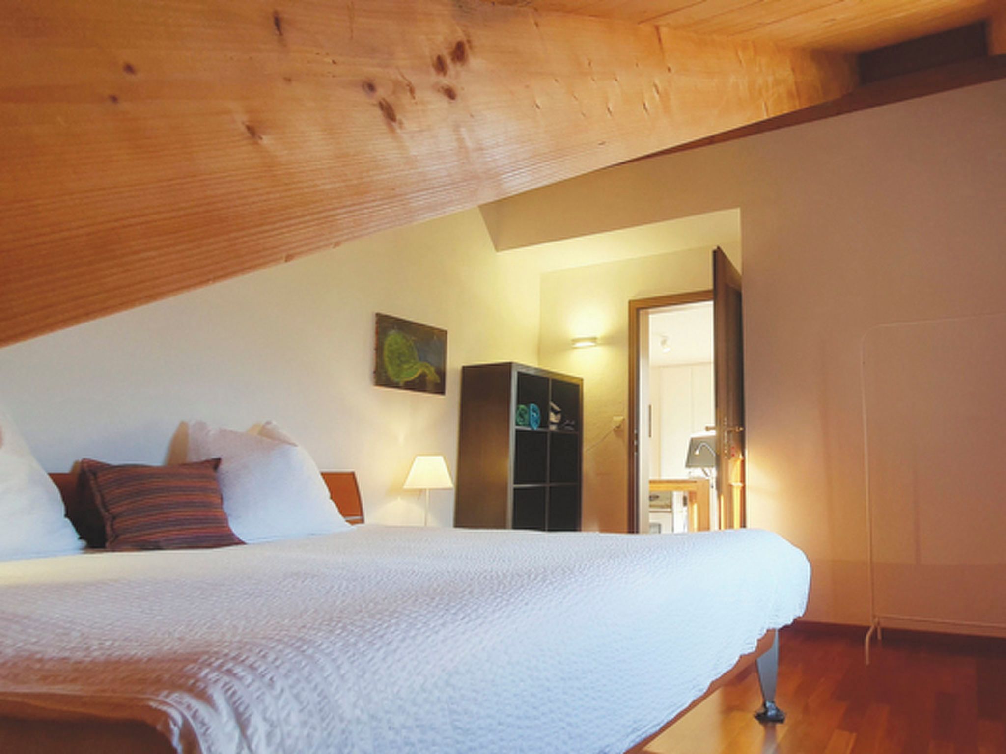 Ein Schlafzimmer mit schrägen Wänden, einem Bett mit weißer Bettwäsche, einem Nachttisch, einer Lampe und einem Kleiderschrank.