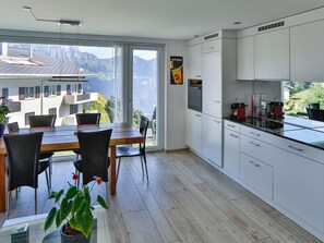 A modern kitchen with white cabinets, a dining area with a wooden table and chairs, and a large window offering a mountain view.