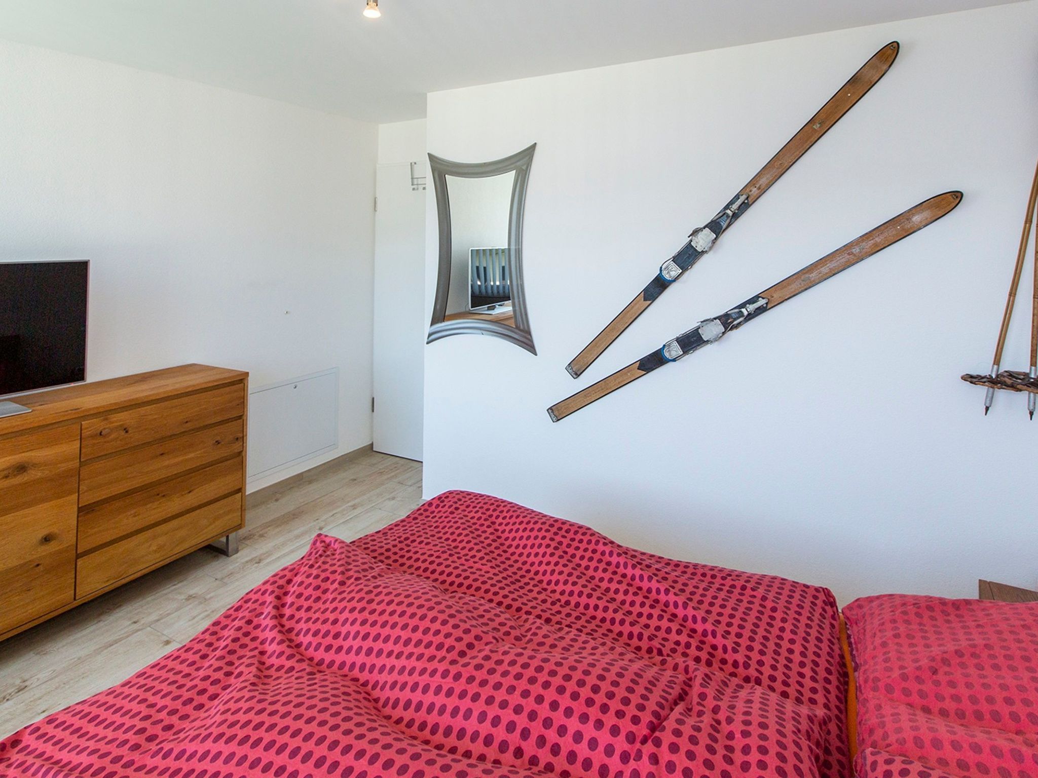 A bedroom with a wooden dresser, a bed with a red polka dot bedspread, a mirror, and skis mounted on the wall.