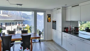Ett modernt kök med vita skåp, en matplats med ett träbord och stolar samt ett stort fönster med utsikt över bergen.
