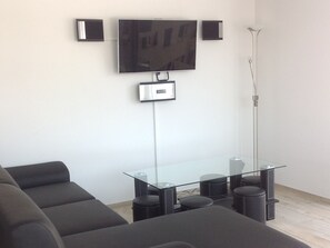 A modern living room with a flat-screen TV mounted on the wall, a glass-top coffee table, black sofas, and a standing lamp.