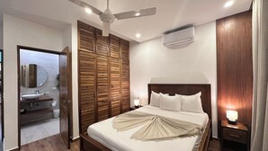 Een hotelkamer met een bed, een plafondventilator, een airconditioner, een nachtkastje met een lamp en een houten garderobe.