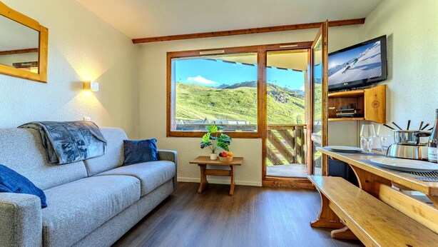 Een gezellige kamer met een bank, een klein tafeltje met een plant, een houten bankje, een televisie en uitzicht op een bergachtig landschap.