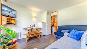 Een ruimtelijk ingerichte leefruimte met kitchenette, een eethoek en een zithoek met een bank en kussens.