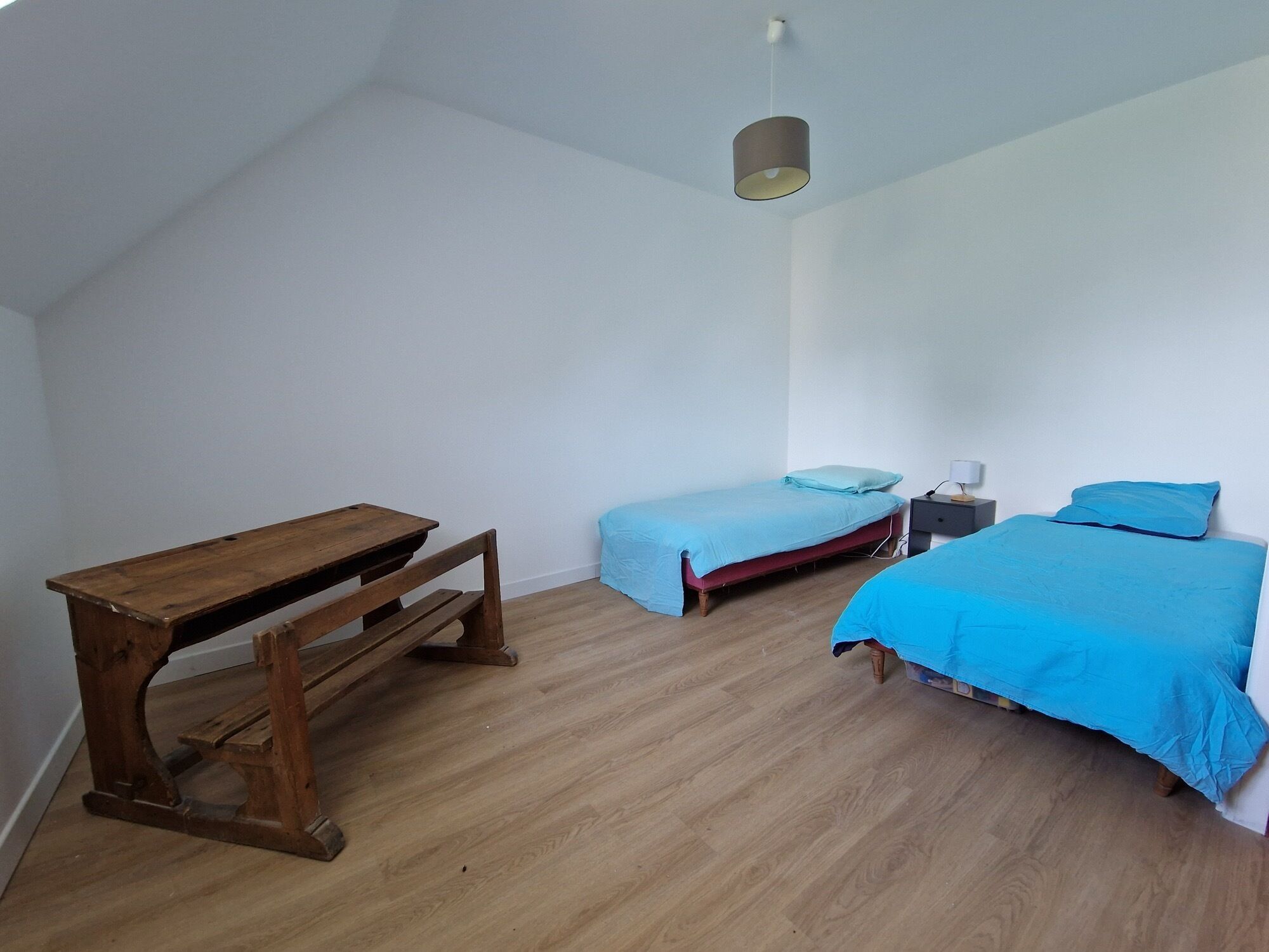 A room with a sloped ceiling, a wooden desk, two single beds with blue bedding, a bedside table with a lamp, and a hanging light fixture.