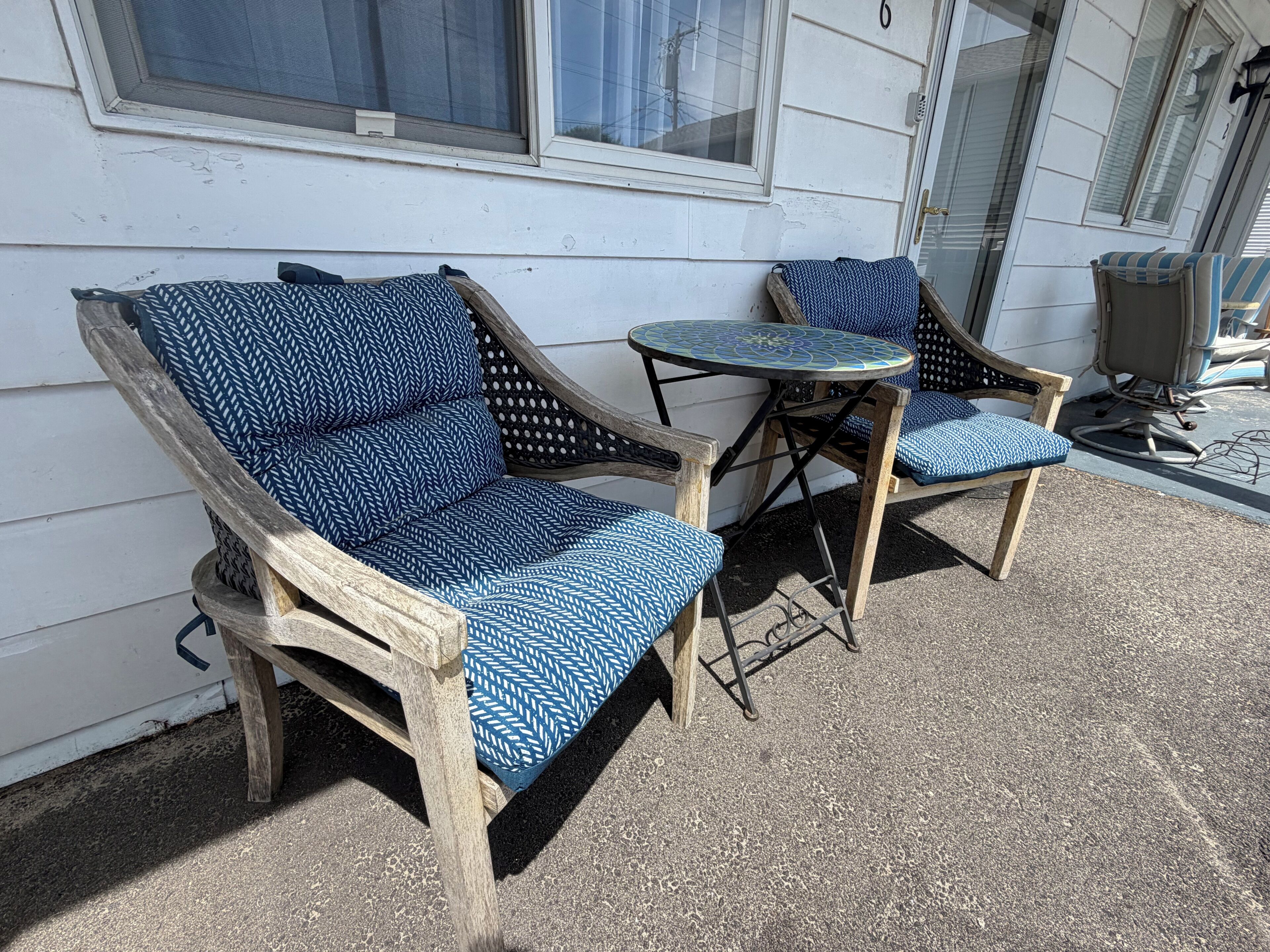 Een terras met twee rieten stoelen en een kleine ronde tafel, elk met blauwe kussens met een patroon.