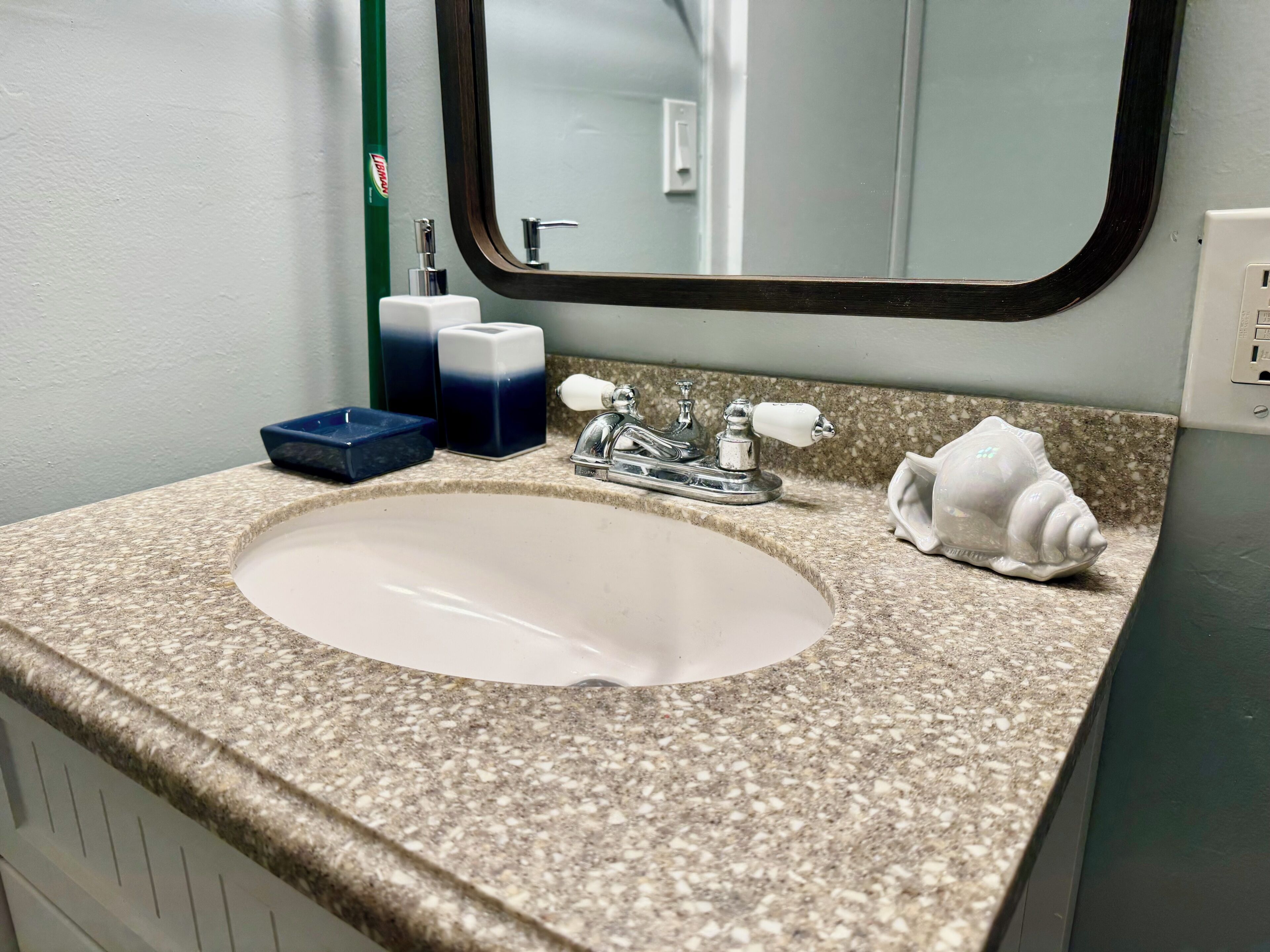 Un baño con encimera de granito, lavamanos blanco, espejo, dispensador de jabón y toallero.