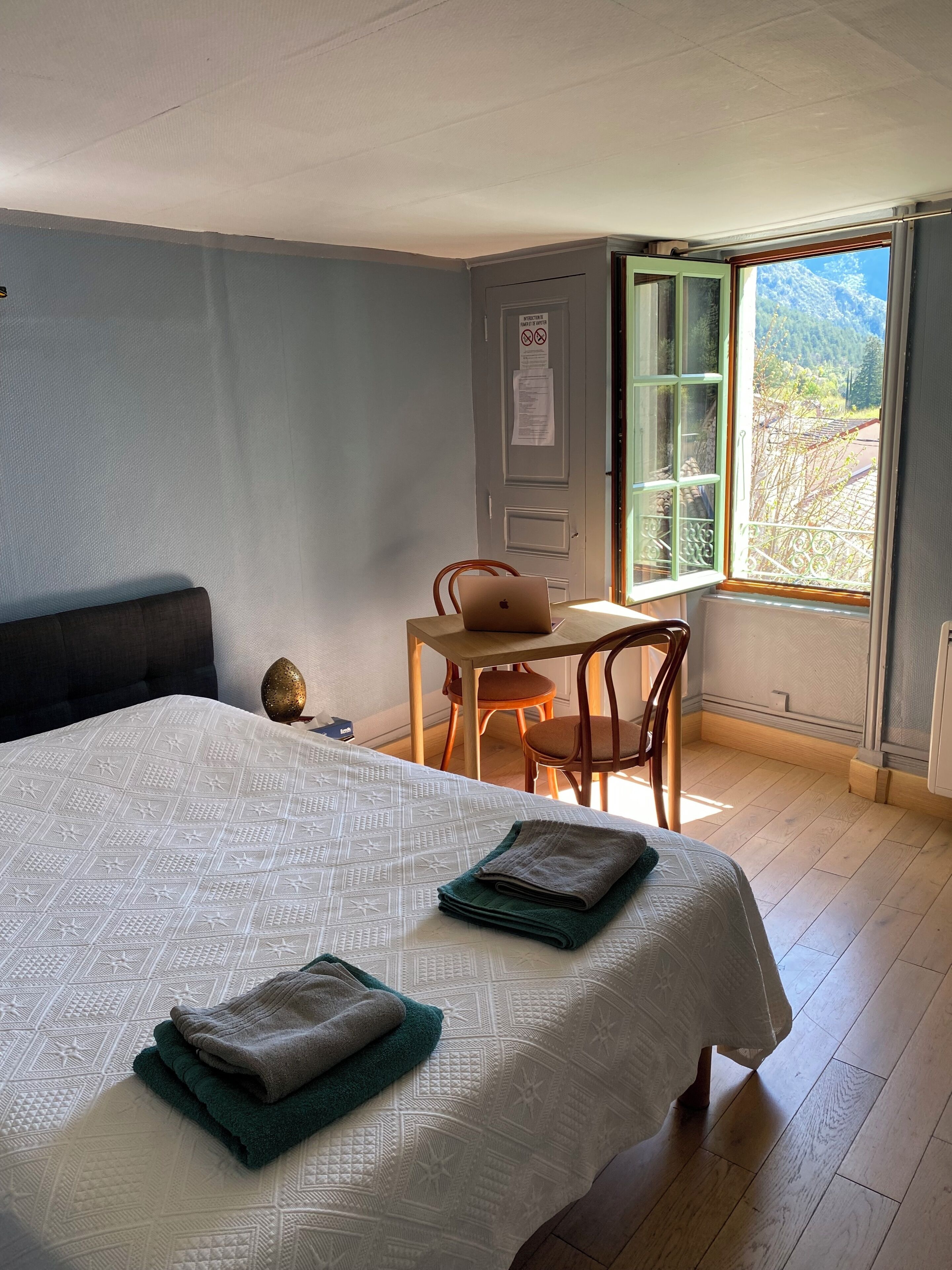 A bedroom with a bed, a desk, and a chair. There is a window with a view of trees and mountains.