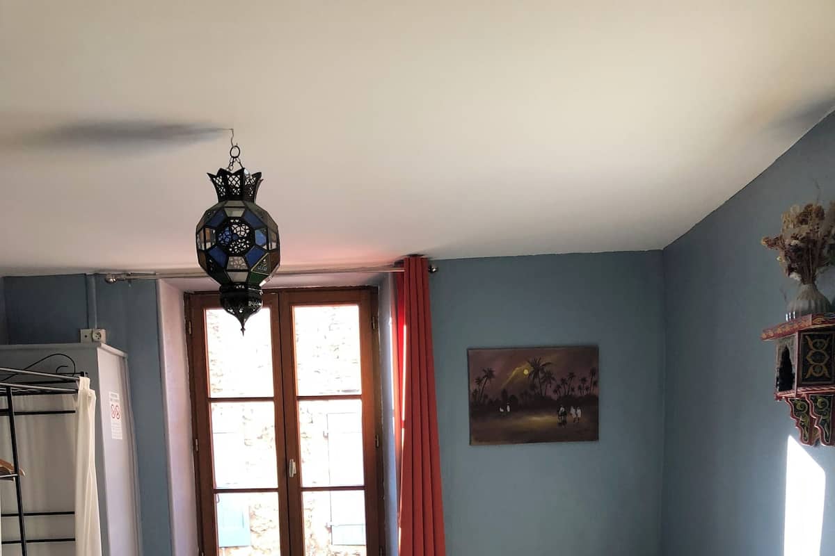 A bedroom with a bed, a radiator, a window with curtains, a lamp, and a painting on the wall.
