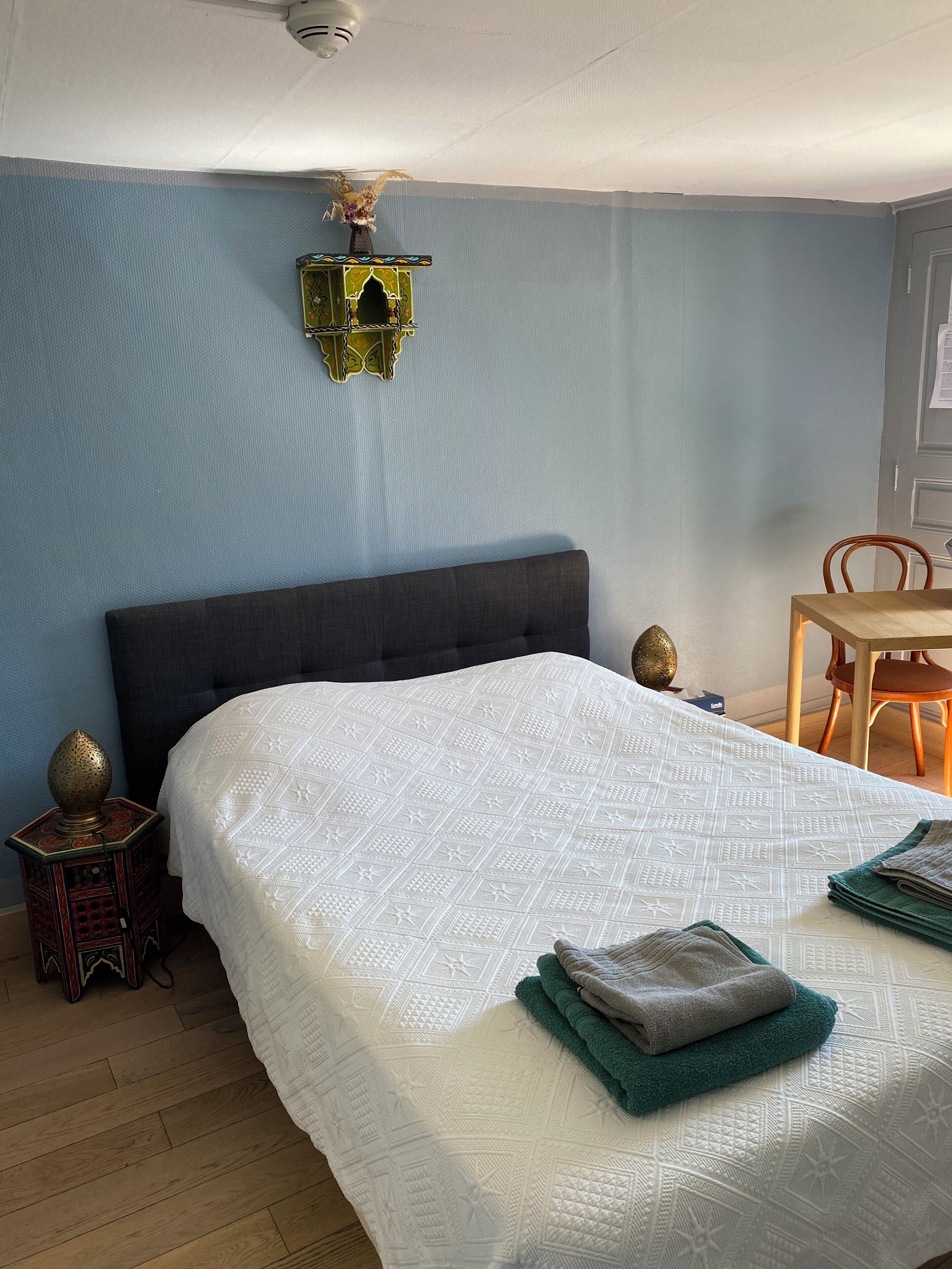 A bedroom with a large bed, a small table, a chair, and a decorative wall piece.
