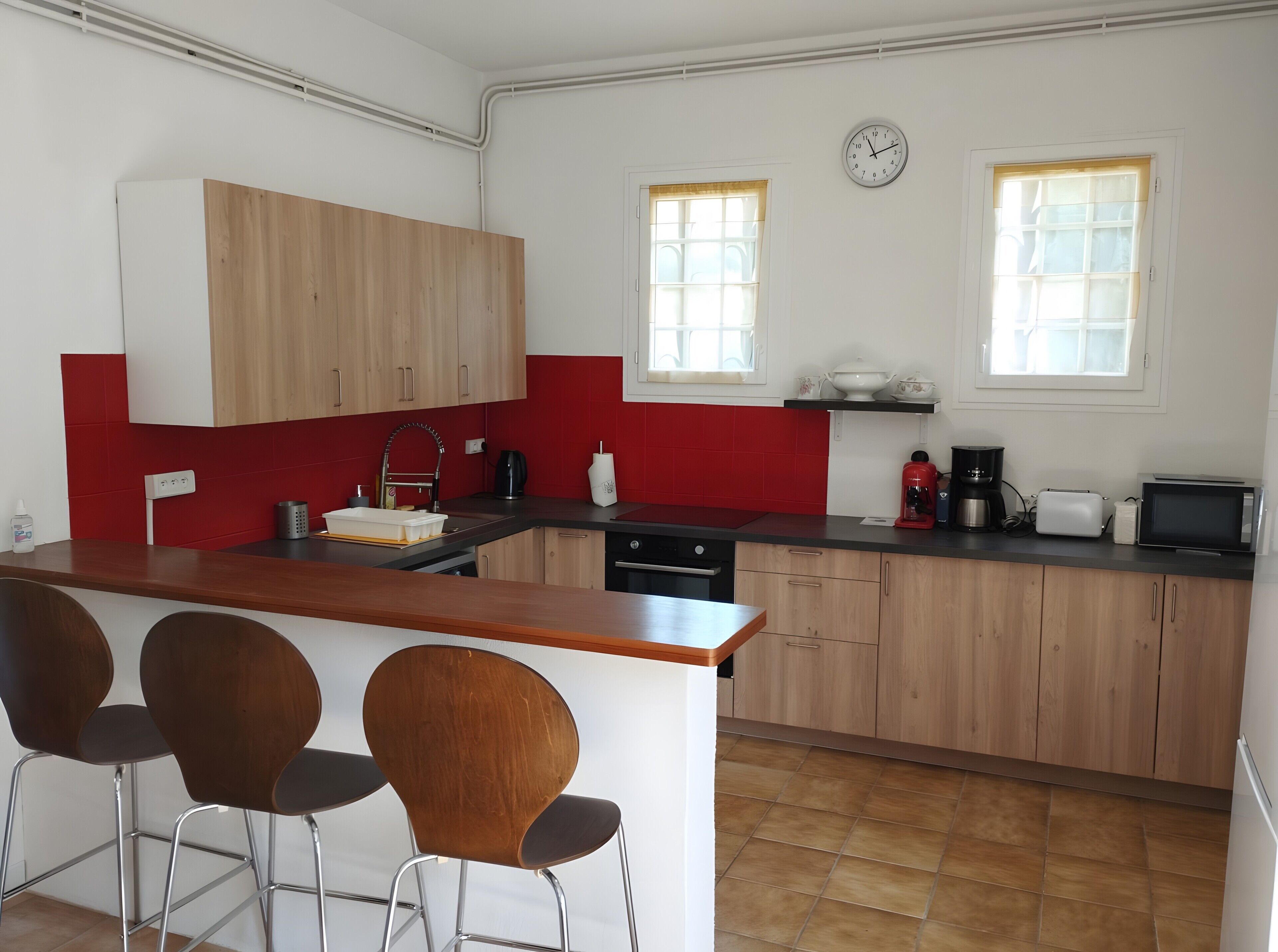 Una cucina con pensili e basi in legno, un lavello, una macchina del caffè, un tostapane e un forno a microonde. Sono presenti due finestre con tende, una sveglia appesa al muro e un'area bar con sgabelli.