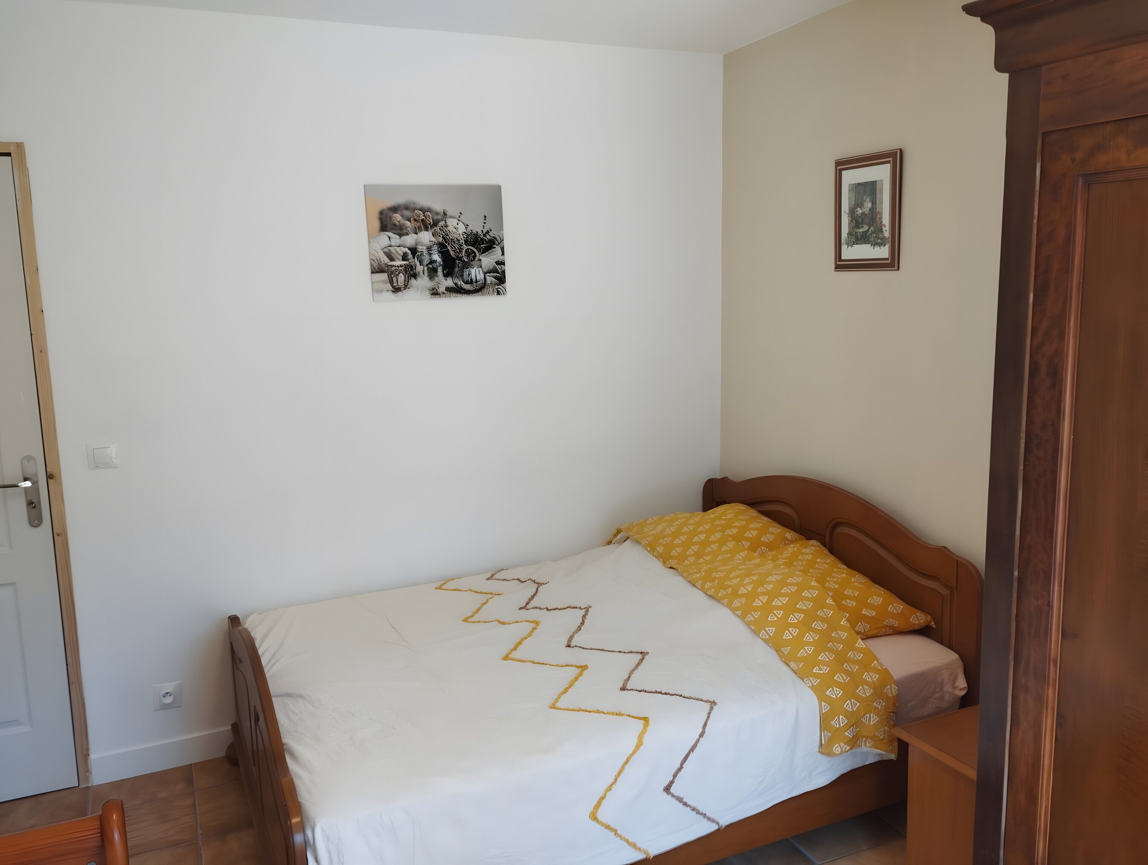 Un dormitorio con una cama, cabecera de madera, pared blanca con un cuadro enmarcado y una puerta.