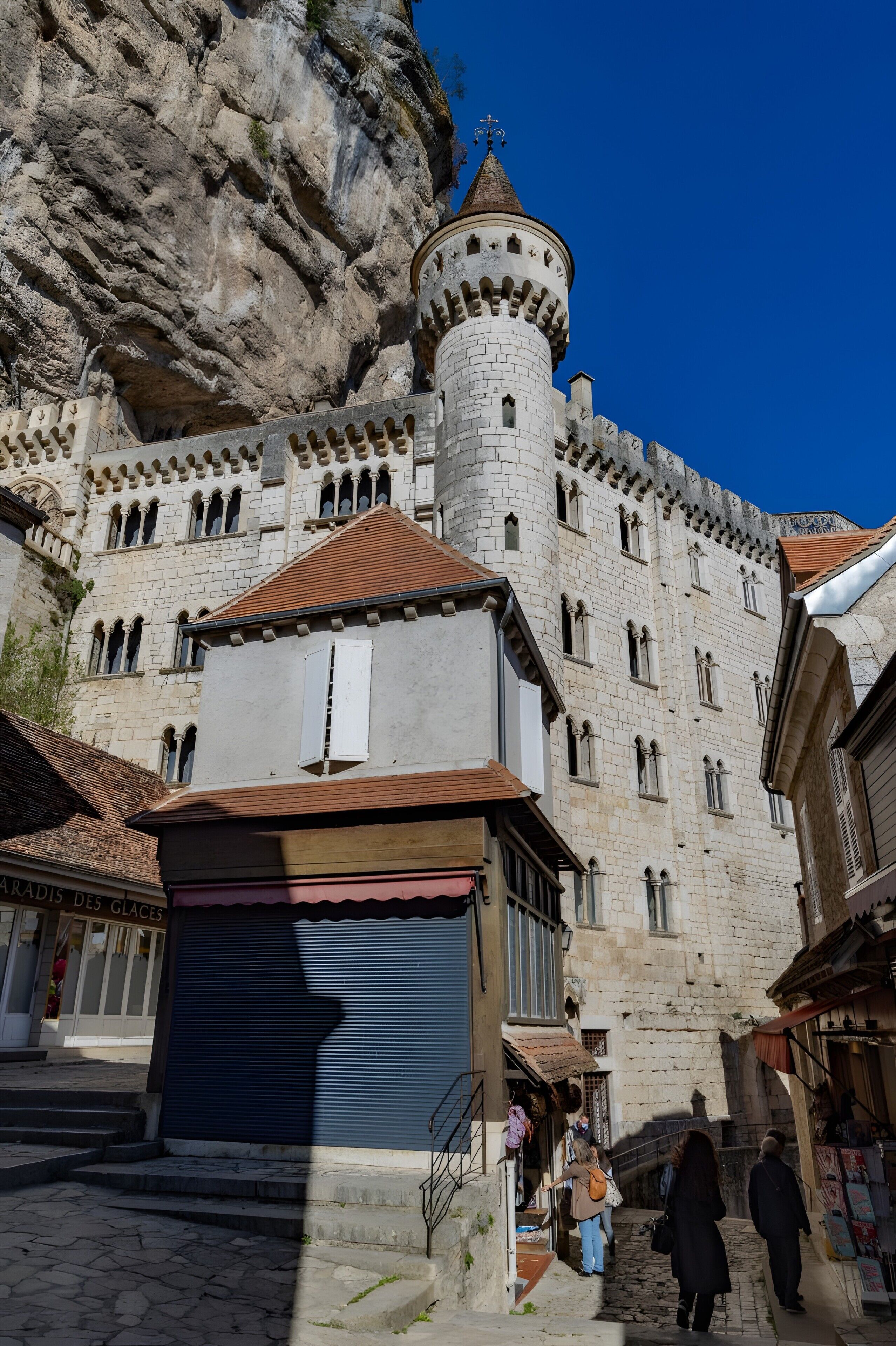 A historic stone building with a tower, a staircase leading to the entrance, and a shop with a blue shutter.