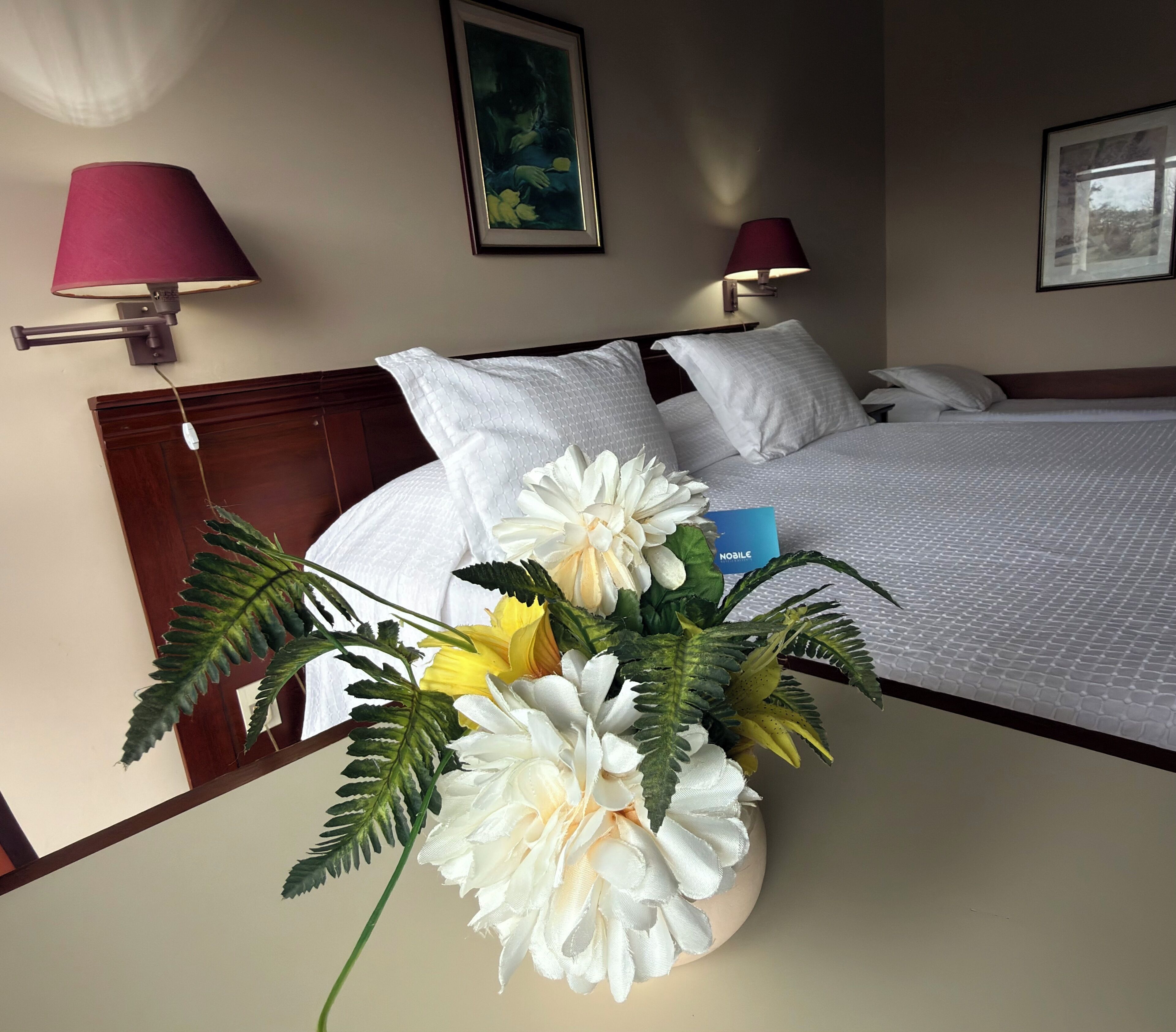 Una habitación de hotel con una cama, dos lámparas de pared, un cuadro enmarcado y un arreglo floral.