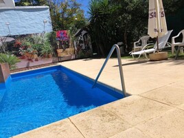Una piscina con escalera metálica, rodeada de un patio con baldosas, sillas reclinables blancas y plantas en macetas.