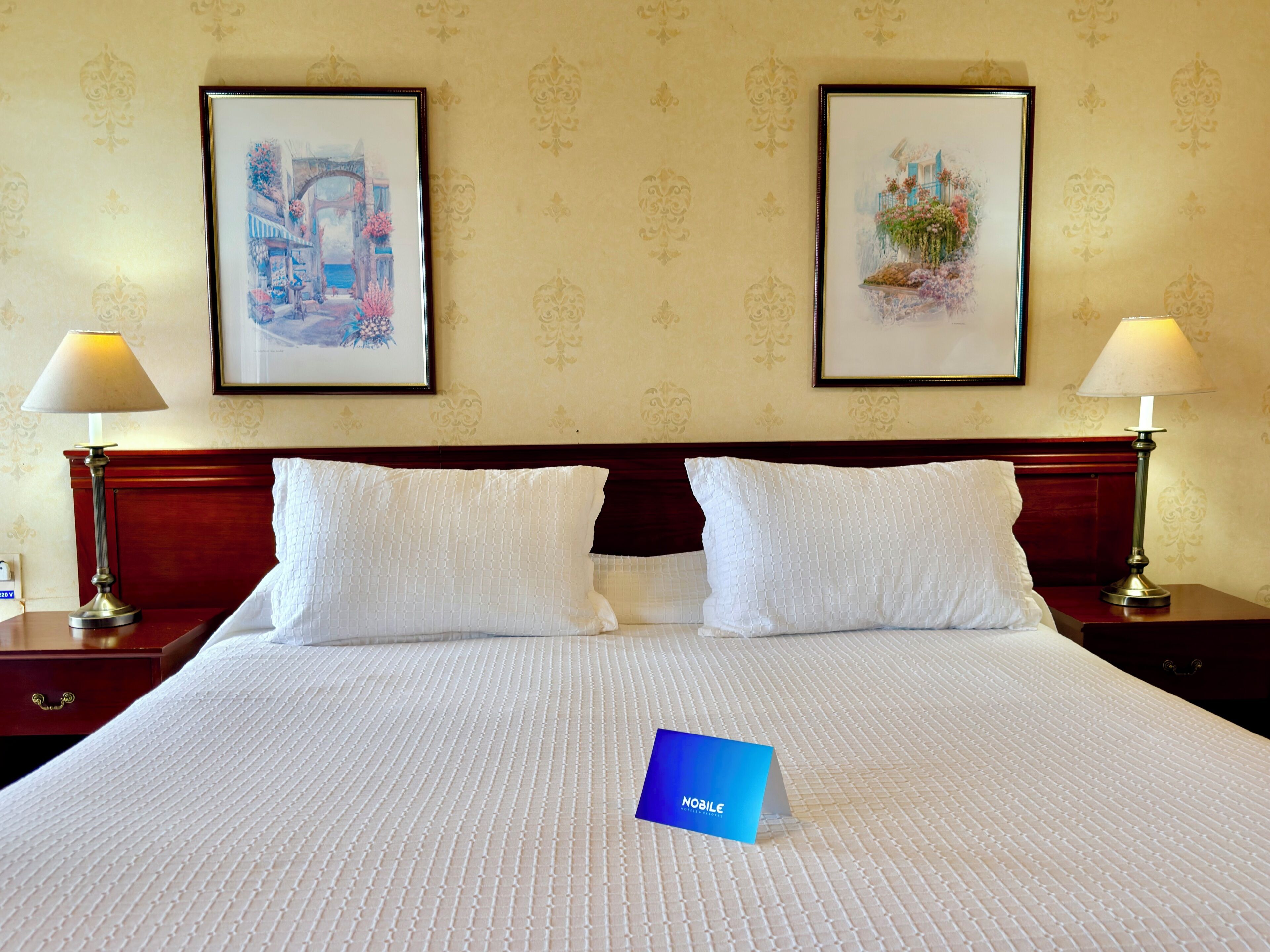 A hotel room with a large bed, two framed artworks on the wall, and two bedside lamps.