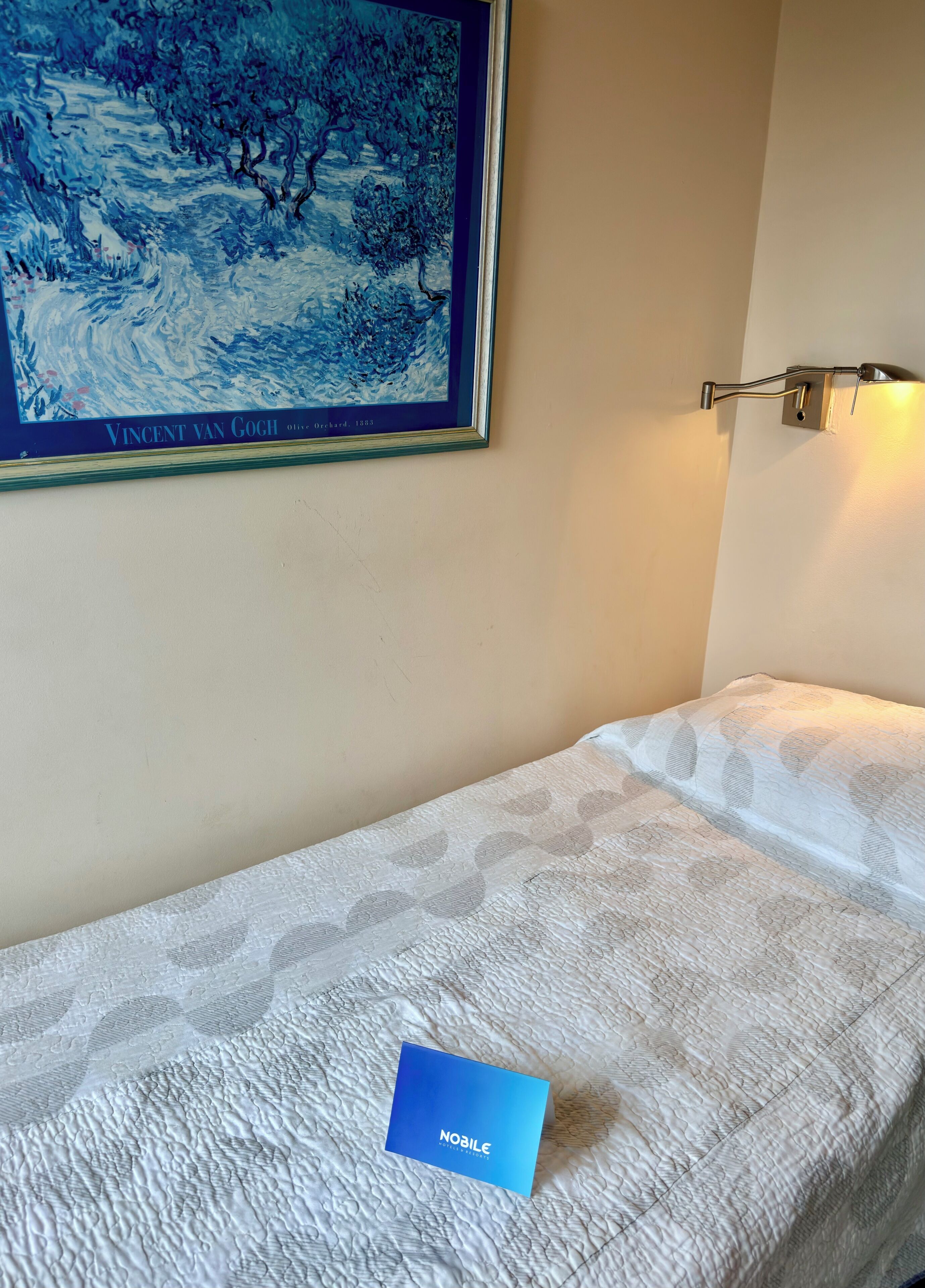 Una cama individual con una colcha blanca texturizada, un cuadro enmarcados de un paisaje azul en la pared y una tarjeta azul pequeña sobre la cama.