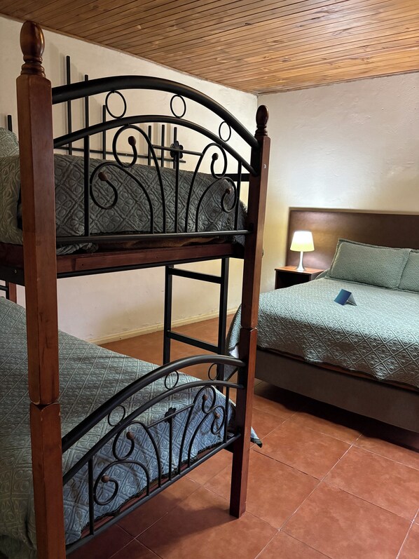 Una cama litera con estructura de madera y cabecera de metal, una mesita de noche con lámpara y una colcha estampada.