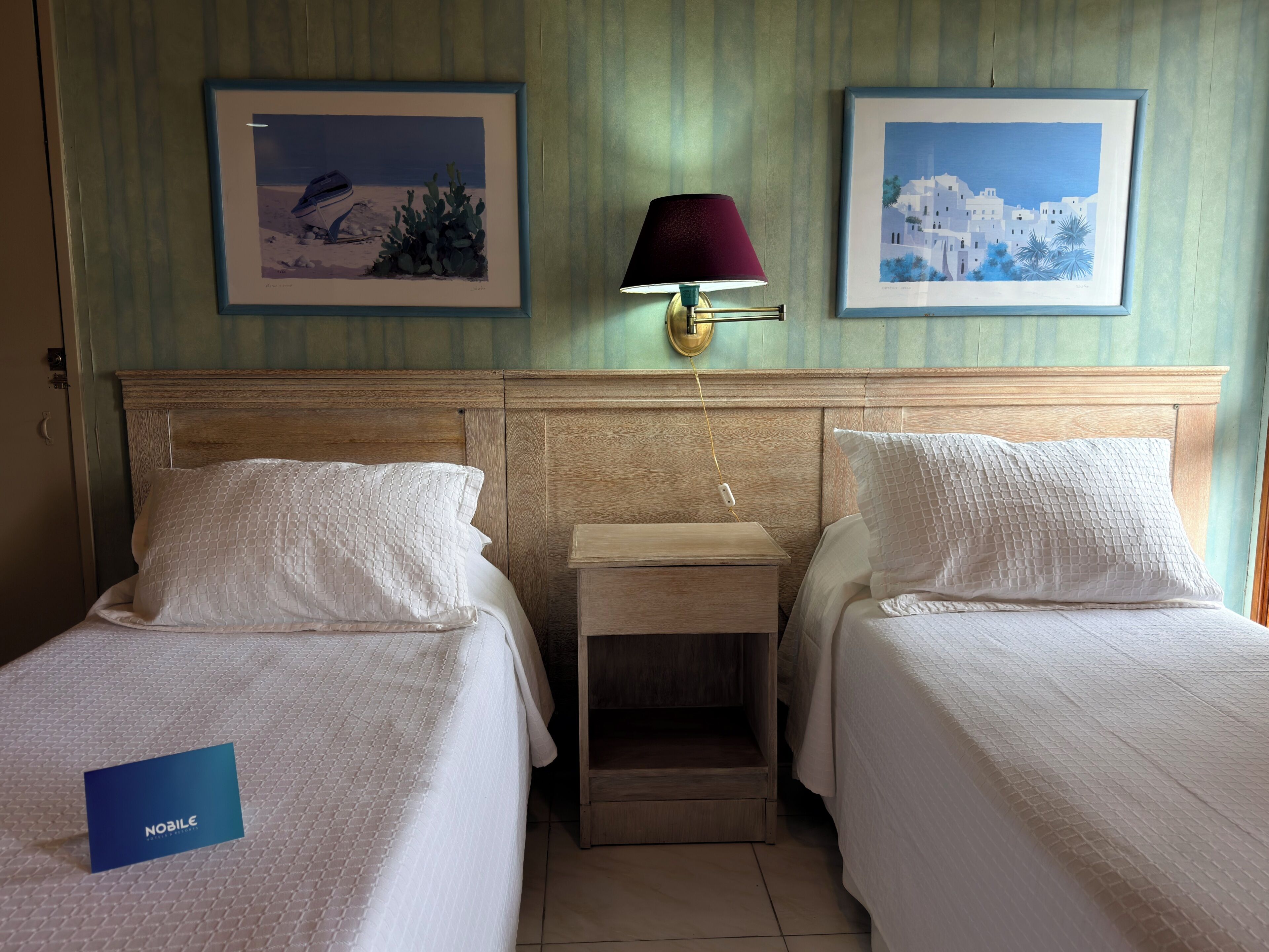 A hotel room with two beds, a nightstand, a lamp, and two framed pictures on the wall.