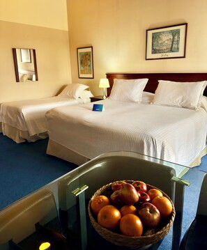 A hotel room with two beds, a bedside table with a lamp, a framed picture on the wall, and a fruit bowl on a glass table.