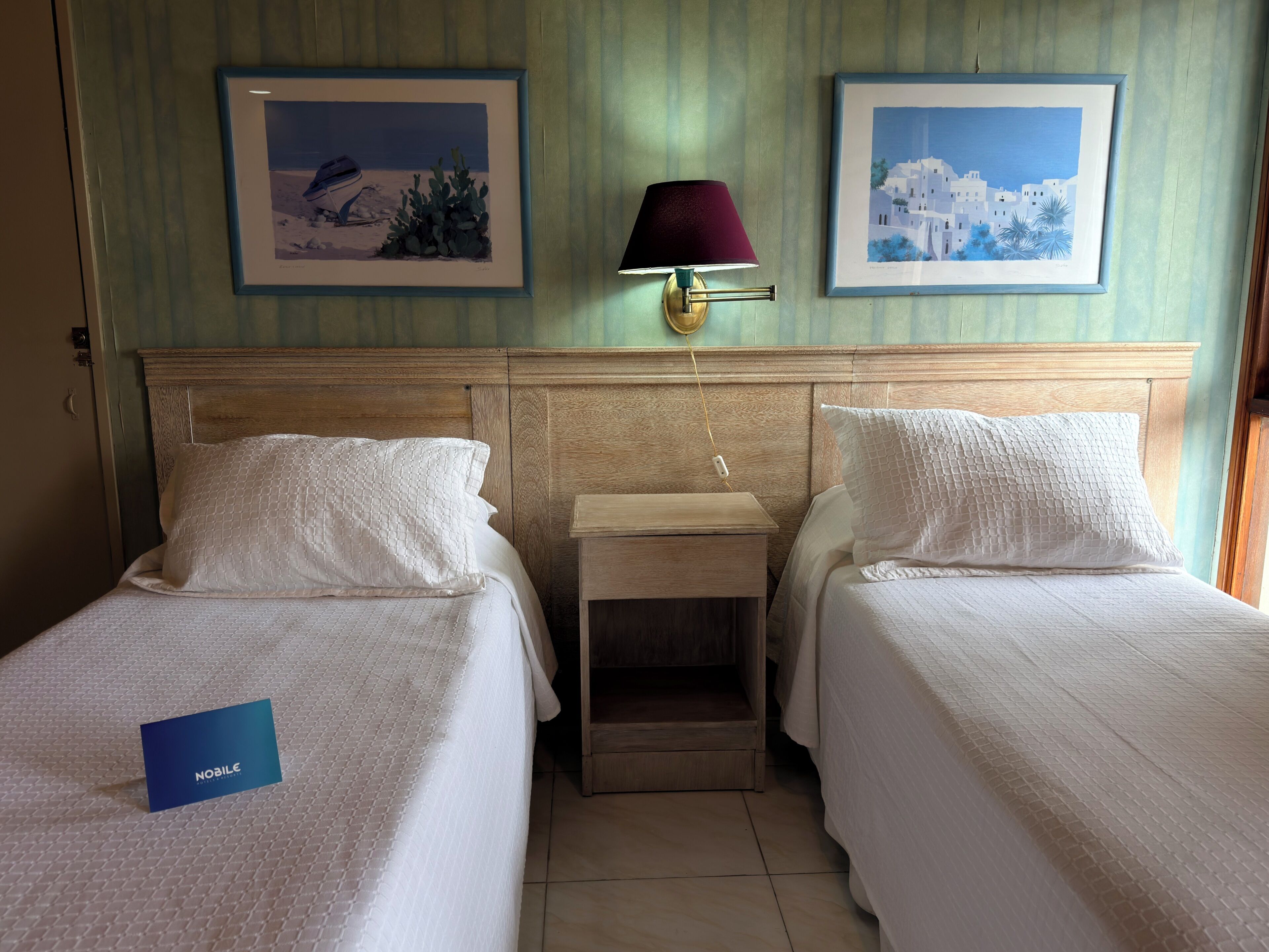 A hotel room with two single beds, a nightstand, a lamp, and two framed pictures on the wall.