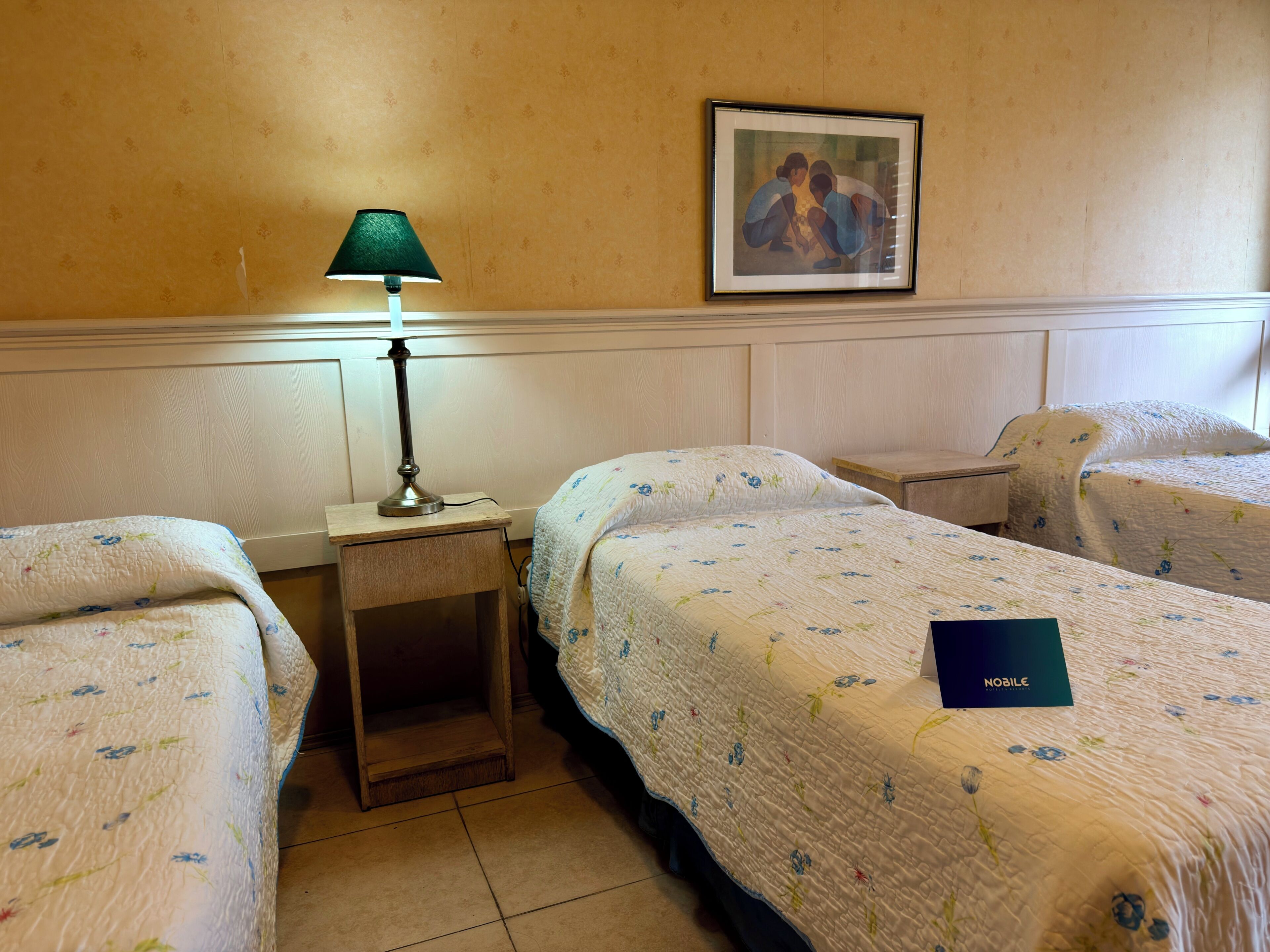 Una camera d'albergo con due letti singoli, un comodino con una lampada, un quadro appeso al muro e un libretto blu su uno dei letti.