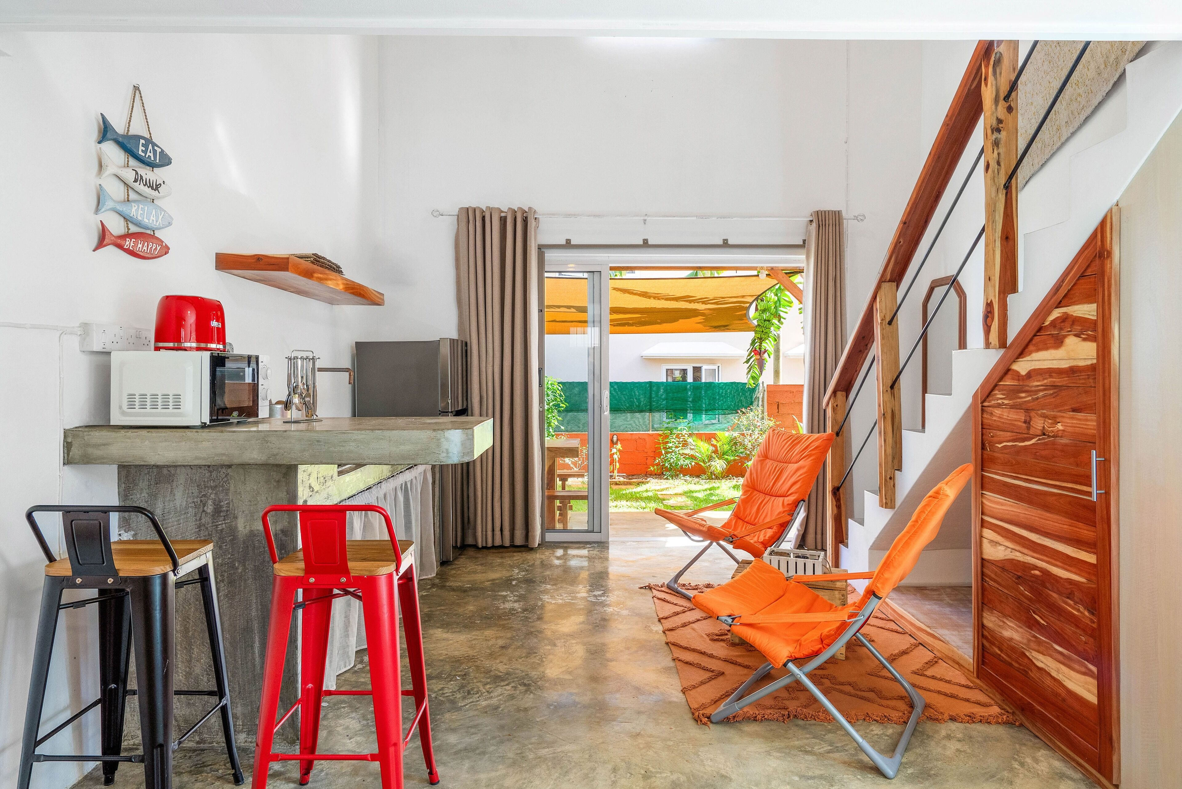 Una cocina compacta con microondas, tostadora y cafetera, una zona de comedor con dos taburetes y vistas a un jardín a través de un ventanal.