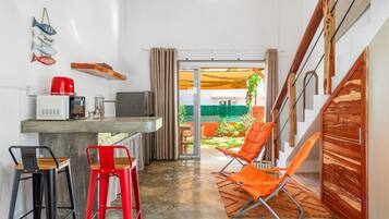 Una cocina compacta con microondas, tostadora y cafetera, una zona de comedor con dos taburetes y vistas a un jardín a través de un ventanal.