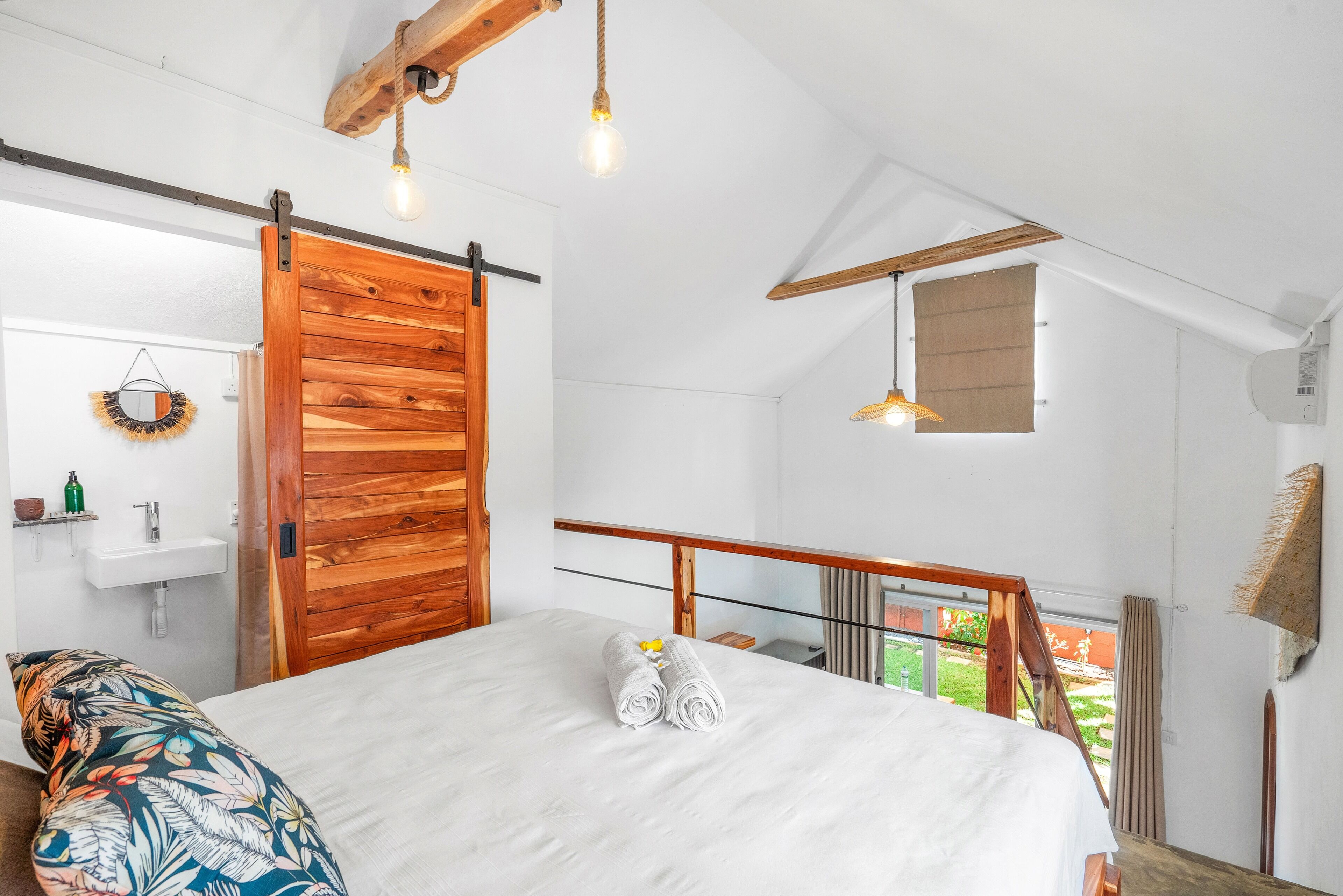 Un dormitorio con puerta de madera, cama con toallas dobladas, lámpara colgante y vistas al exterior.