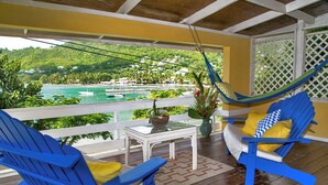 Ein Balkon mit blauen Stühlen, einem weißen Tisch und Blick auf eine Küstenlandschaft mit Gebäuden und Grünflächen.