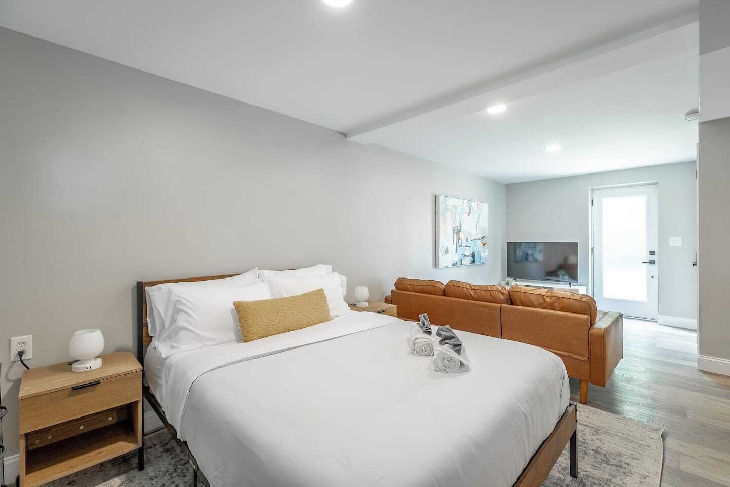 A modern bedroom with a large bed, a wooden nightstand, a TV, and a sofa in the adjacent living area.