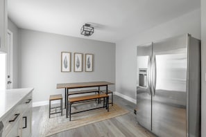 A modern kitchen with a stainless steel refrigerator, a wooden dining table with benches, and three framed leaf artworks on the wall.