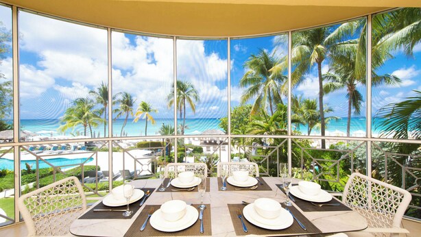 Comedor con una mesa lista para cuatro personas, con vistas a una piscina y palmeras.