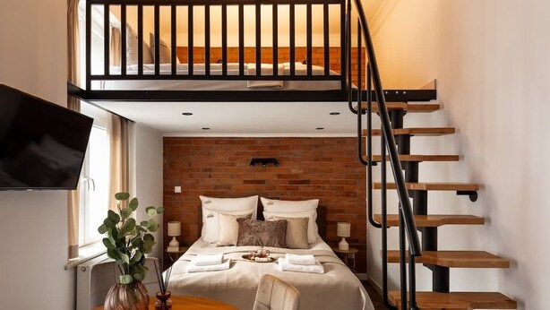 A bedroom with a loft bed, a brick wall, a wooden staircase, a TV, a dining table with chairs, and a vase with plants. - L23 Boutique 18B | City Center (Kraków)