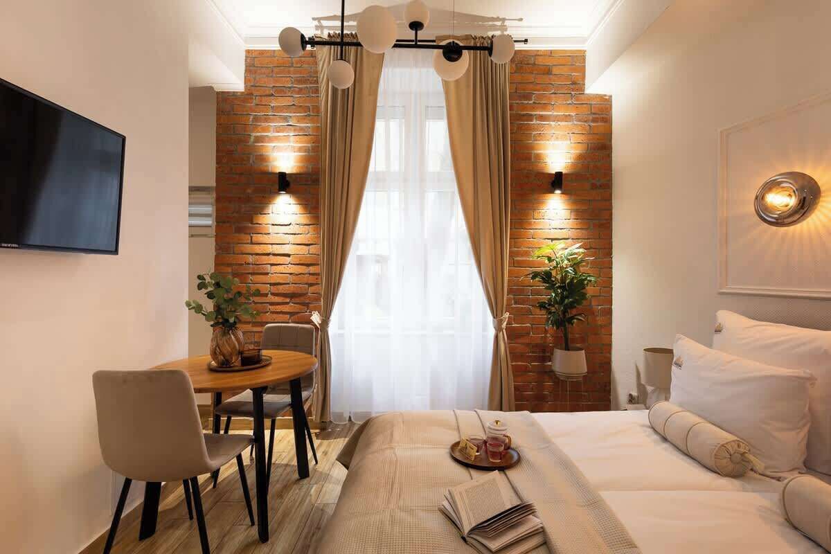 A hotel room with a brick wall, a bed with white bedding, a dining table with chairs, and a TV mounted on the wall.