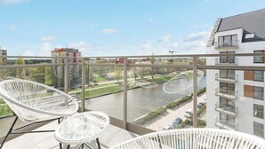 Un balcón con sillas de mimbre blanco y barandilla de vidrio con vistas a un río y edificios.