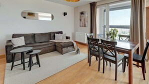Ein modernes Wohnzimmer mit Ecksofa, Esstisch mit Stühlen, einem kleinen Couchtisch, einem großen Fenster mit Vorhängen und einer Deckenleuchte.