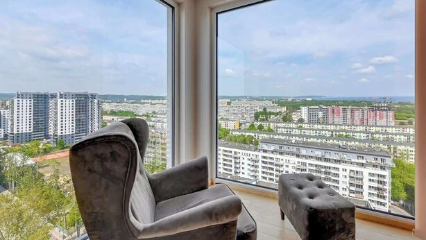 Ein Zimmer mit einem großen Fenster, einem grauen Sessel und einem kleinen Hocker.