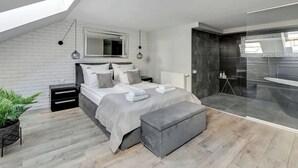 Quarto moderno com claraboia, cama grande, mesa de cabeceira, planta e área de chuveiro em vidro.